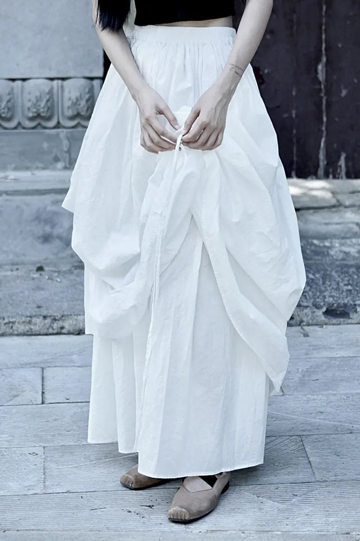 Avant-garde off-white cotton maxi skirt with dramatic asymmetrical draping and an adjustable drawstring hem, shown on a model in an outdoor setting.