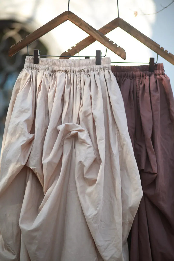 Two Kylori avant-garde maxi skirts hanging on hangers, showcasing a voluminous asymmetrical draped silhouette in off-white and muted mauve cotton.