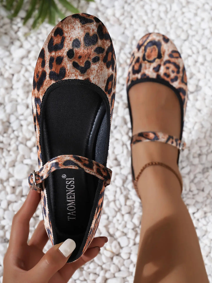 A pair of leopard print Mary Jane ballet flats, one held by a hand revealing the insole, the other worn on a leg with an ankle strap and buckle, set against a white pebble background.