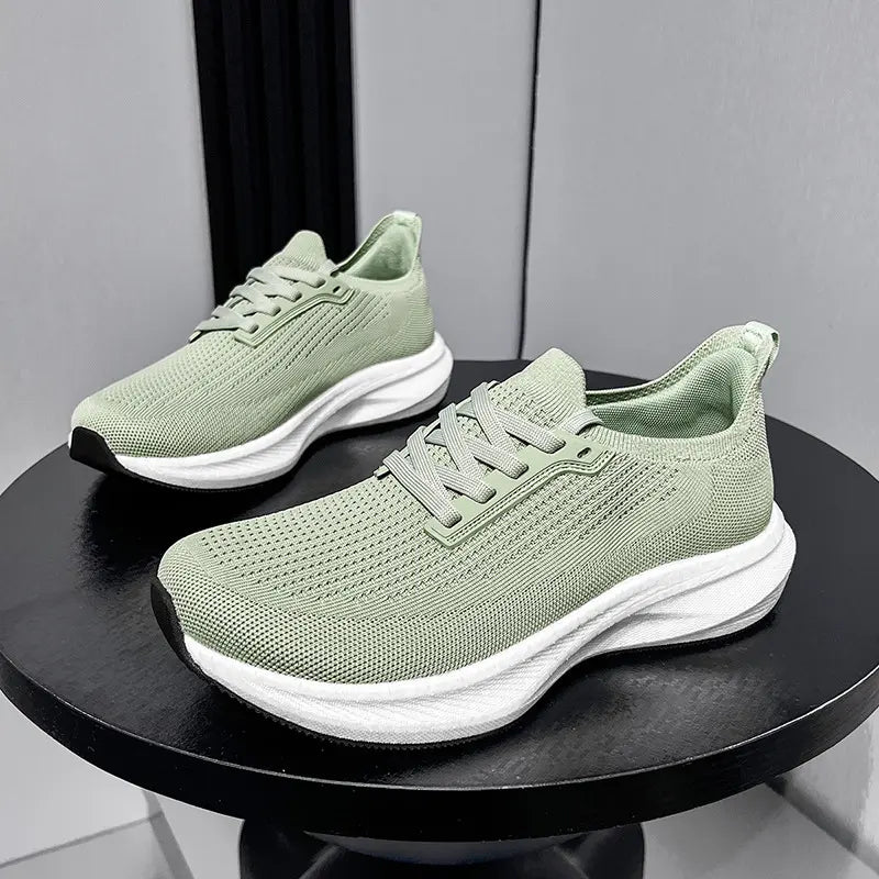 A pair of light green wide-fit breathable athletic trainers shown from a three-quarter side view on a dark circular table against a grey background.
