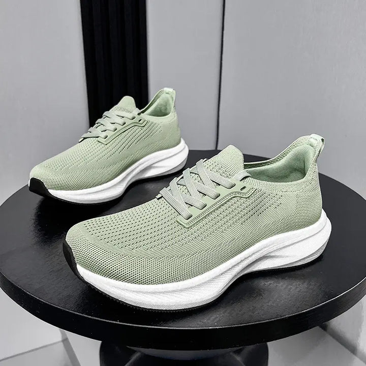 A pair of light green wide-fit breathable athletic trainers shown from a three-quarter side view on a dark circular table against a grey background.