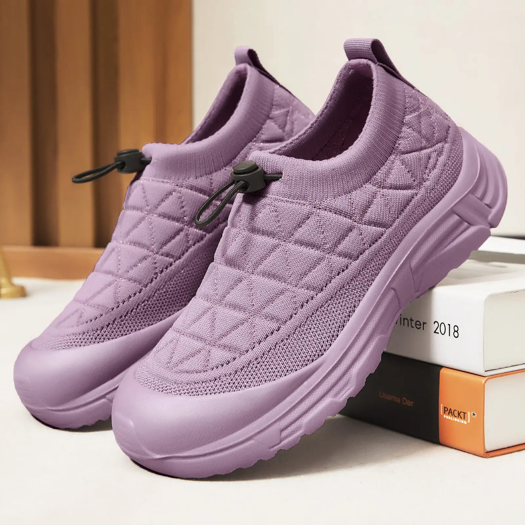 Pastel lilac breathable mesh slip-on sneakers featuring a quilted knit upper, bungee laces, and a thick white platform sole, presented from a three-quarter angle on a studio surface with decorative books.