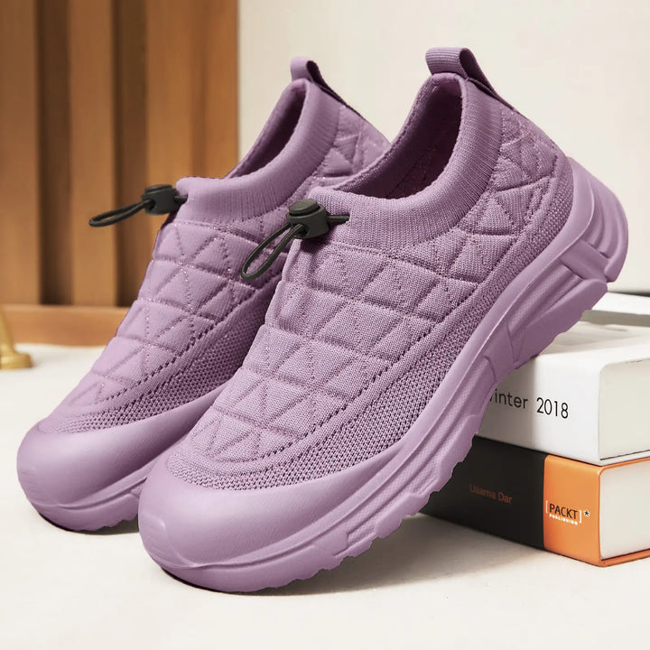 Pastel lilac breathable mesh slip-on sneakers featuring a quilted knit upper, bungee laces, and a thick white platform sole, presented from a three-quarter angle on a studio surface with decorative books.