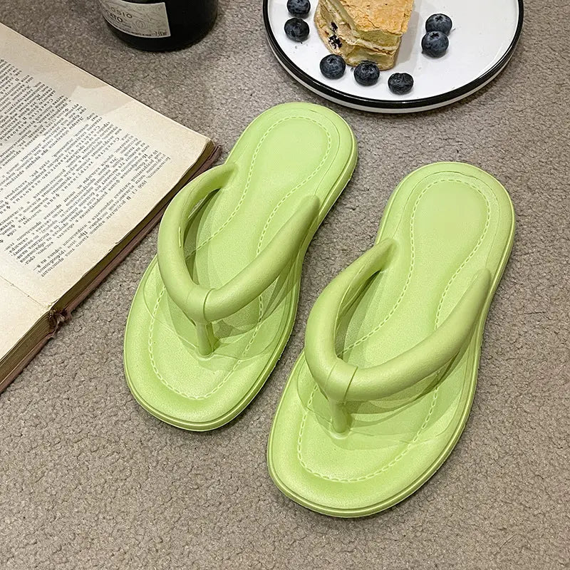 Light green EVA flip-flops with a cushioned thong strap, styled on a textured surface next to an open book and a plate of food.