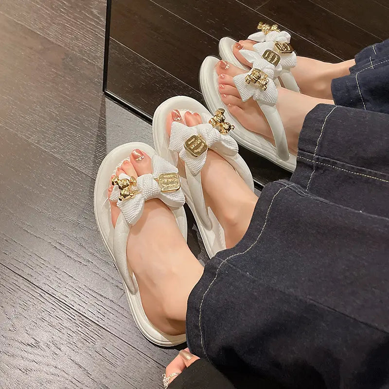 A pair of white Lyraku flip-flops with white textured bows and gold teddy bear charms are worn by a person on a wooden floor, showcasing their playful summer style.