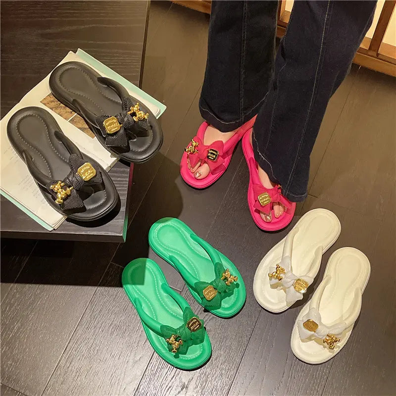 Women's Lyraku flip-flops in multiple colors, featuring a bow and gold teddy bear charm, shown in a flat lay arrangement with legs wearing pink ones.
