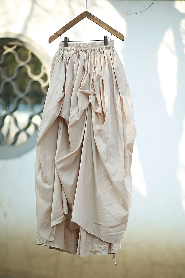 Voluminous off-white cotton maxi skirt with dramatic asymmetrical draping and an adjustable drawstring hem, displayed hanging from a wooden hanger.