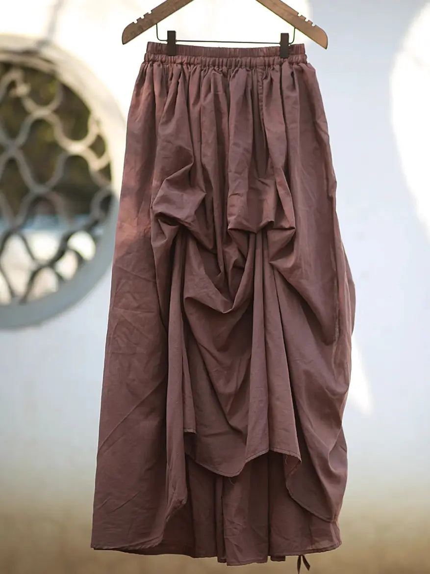 Avant-garde asymmetrical draped maxi skirt in muted rose cotton, hanging from a wooden hanger with an adjustable drawstring hem and elasticated waistband.