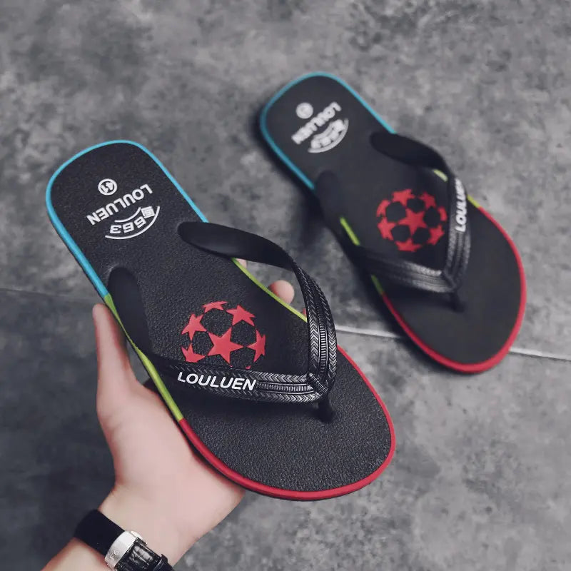 Pair of black men's rubber flip-flops with textured straps, a red star graphic, and a multi-colored block sole featuring red, green, and blue accents. One flip-flop is held by a hand.