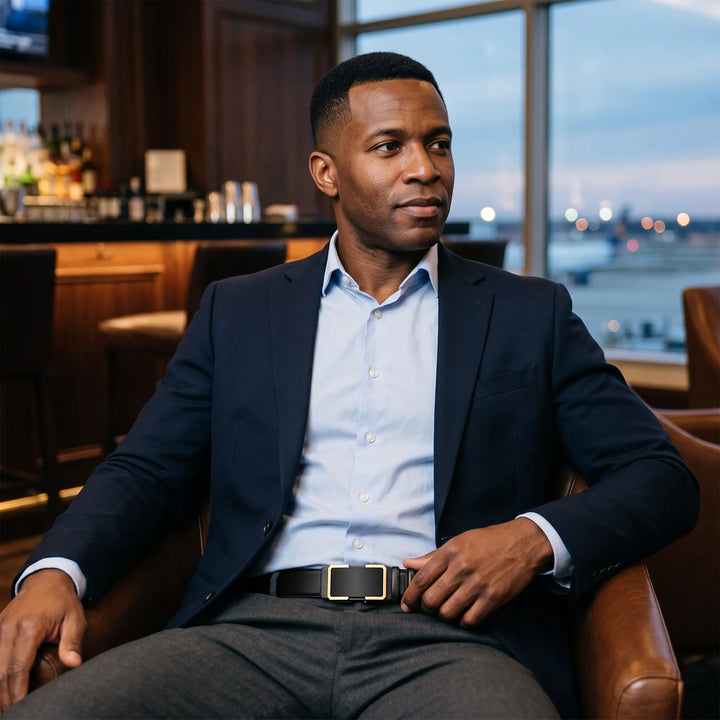 A man wearing a navy suit jacket and grey trousers sports a Zentir black leather automatic belt with a matte black buckle featuring a sophisticated gold-toned frame, seated in a stylish lounge.