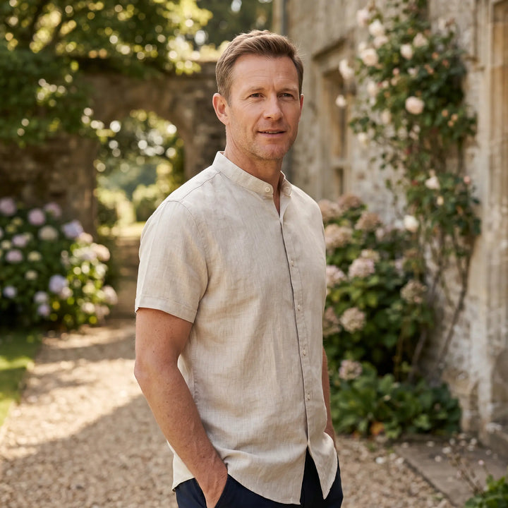 A man wearing a light beige short-sleeve linen blend shirt with a mandarin collar, standing outdoors in a picturesque garden setting.