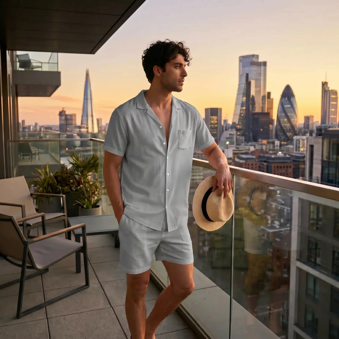 Men's relaxed-fit soft grey linen blend set featuring a camp collar shirt and mid-thigh shorts, shown on a model on a city balcony at sunset.