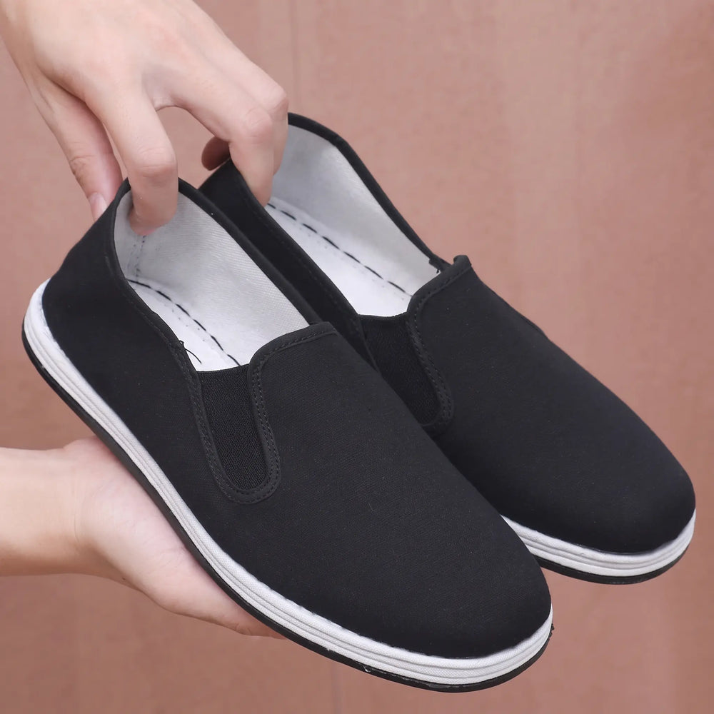 Men's black canvas slip-on loafers held by hands, showing the round toe, elasticated side panels, and white rubber sole stripe. Features traditional cloth shoe construction and a textured brown background.