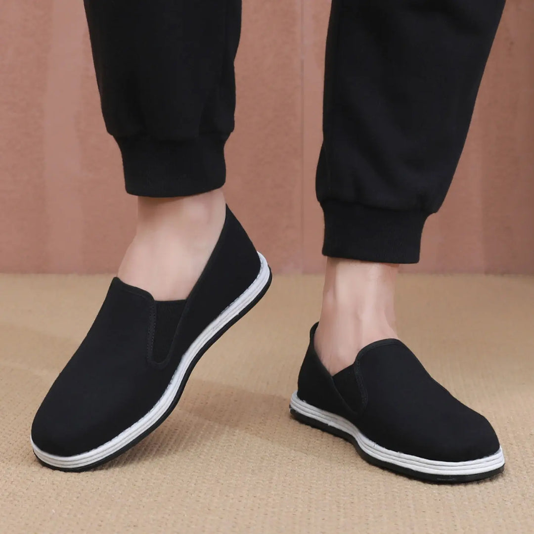 Men's black canvas slip-on shoes worn casually, featuring a rounded toe, elasticated side panels, and a white rubber stripe on the sole. Shown from a three-quarter view on a neutral textured surface.