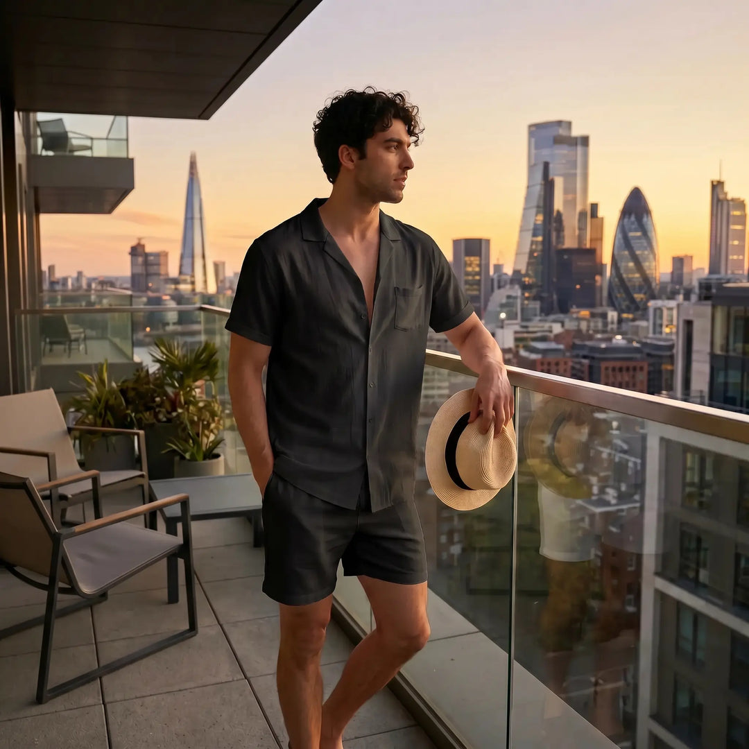 A man wearing a black linen blend relaxed-fit two-piece set, featuring a camp collar shirt and matching shorts, stands on a balcony overlooking a city skyline at sunset.