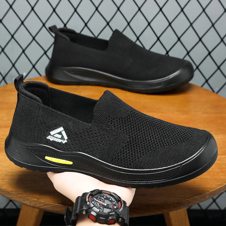 Black breathable mesh slip-on sneakers for men, shown in a three-quarter view, featuring a charcoal grey polyurethane sole with a subtle yellow accent and a white triangular logo.