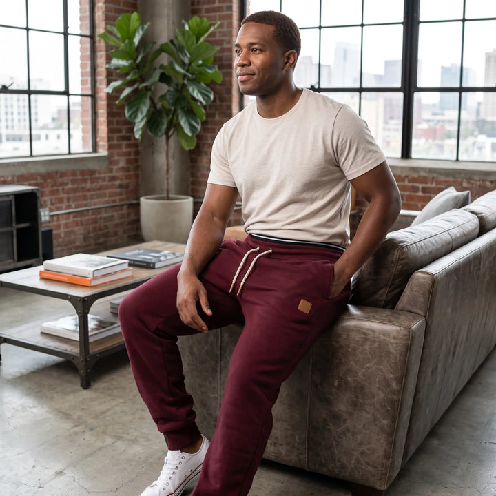 A man wearing burgundy fleece-lined tapered joggers and a light t-shirt, seated casually on a leather couch in a modern loft living space.