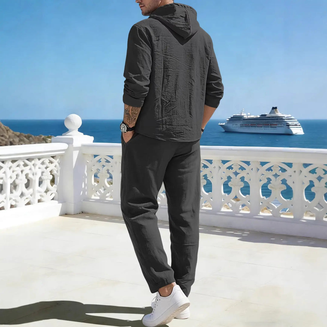 Man from behind wearing a dark grey crinkled linen blend hooded tracksuit set, standing on a balcony overlooking the sea with a cruise ship.