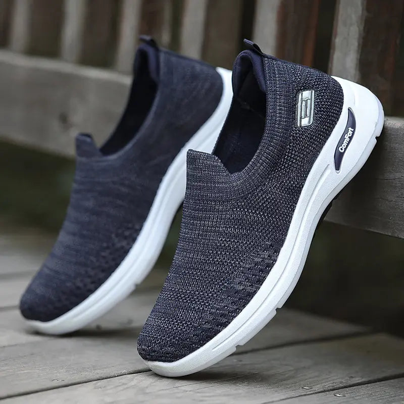 A pair of men's dark grey marl knit slip-on sneakers shown at a three-quarter angle, featuring a white midsole with navy blue 'Comfort' branding and a black rubber outsole.