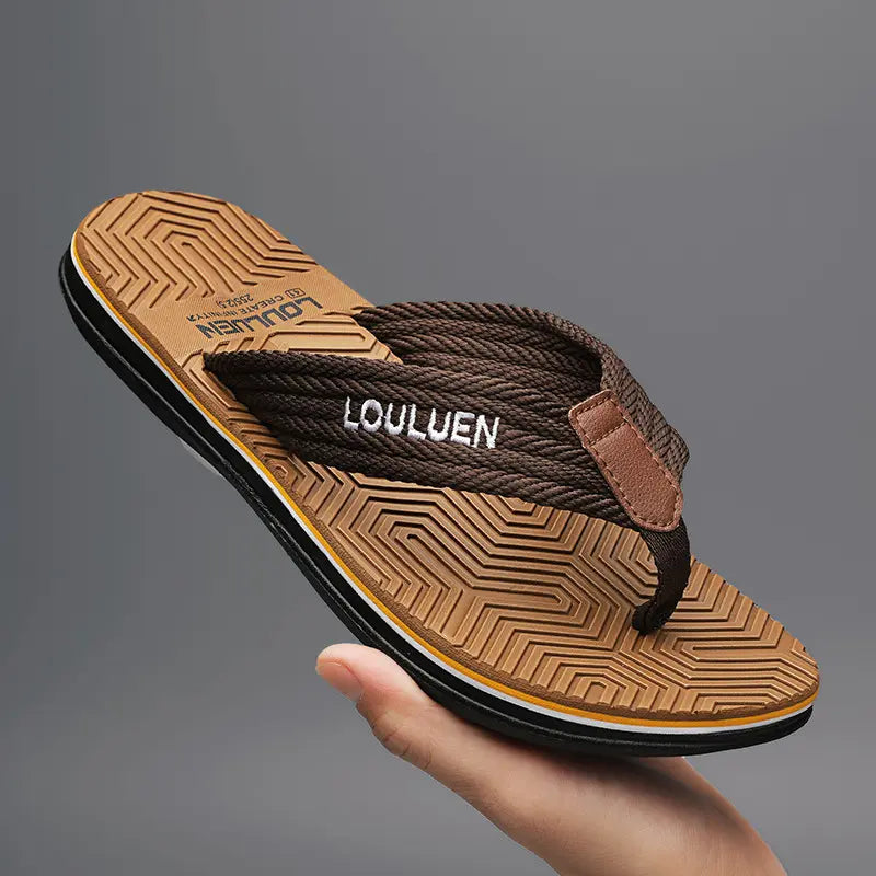 Men's flip-flops with a textured brown braided fabric strap featuring white embroidered 'LOULUEN' text, set against a molded EVA footbed with a geometric pattern, held in hand on a grey studio background.