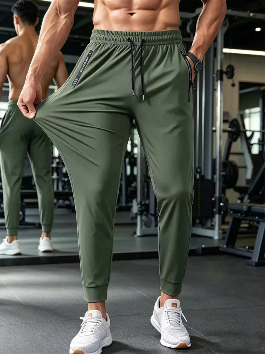 Men's forest green lightweight and high-stretch jogger trousers shown on a model in a gym, featuring an elasticated drawstring waist and zippered pockets.