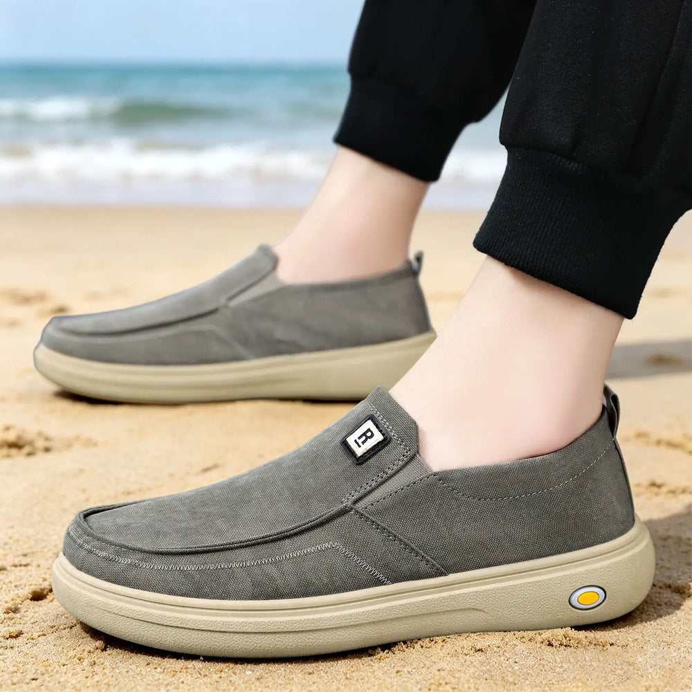 Men's Rhylo grey canvas slip-on loafers shown worn on the feet on a sandy beach, featuring a lightweight breathable design and a light tan rubber sole.