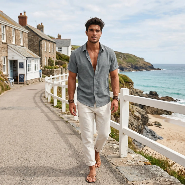 A man walks along a seaside promenade wearing a medium grey linen-blend short-sleeve shirt, unbuttoned at the collar, paired with relaxed white pants and sandals, conveying effortless summer style.