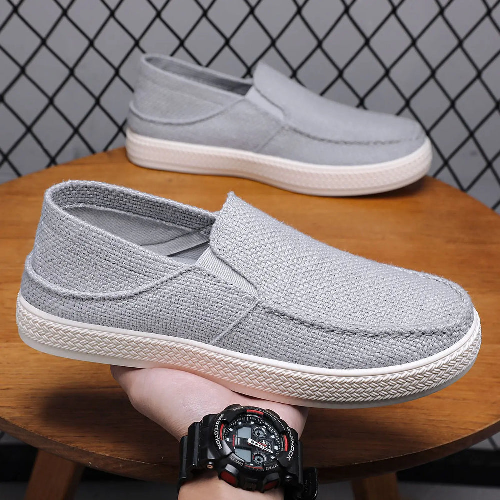 Grey men's breathable mesh slip-on sneakers with a white rubber sole, shown from a three-quarter angle being held by a hand.