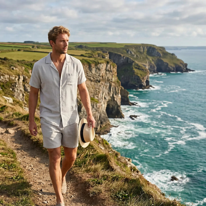Man in a relaxed-fit, light grey linen blend camp collar shirt and matching shorts walking on a scenic coastal path, holding a straw hat.