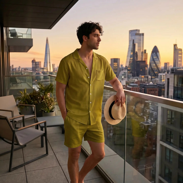 A man wearing a relaxed-fit olive green linen blend short-sleeve camp collar shirt and matching shorts stands on a balcony overlooking a city skyline at sunset, holding a straw hat.