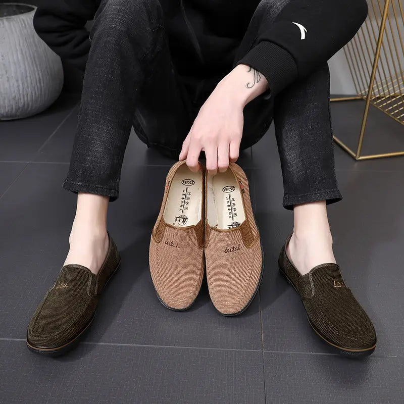 Men's Kryston casual slip-on loafers in dark olive green and warm tan ribbed canvas, shown on-foot with a pair held between the legs.