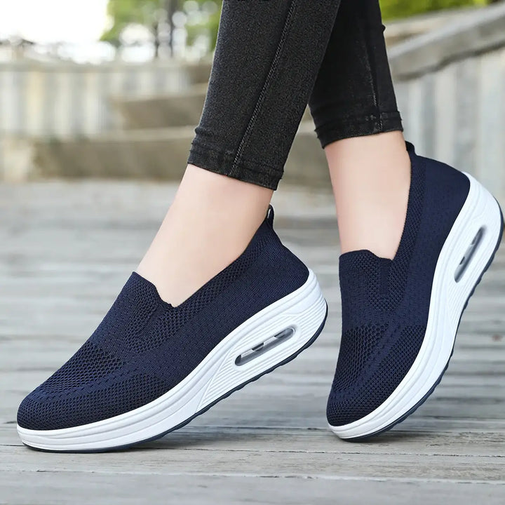 Navy blue breathable knit slip-on sneakers with white platform soles and visible air cushioning, worn by a person on a wooden deck.