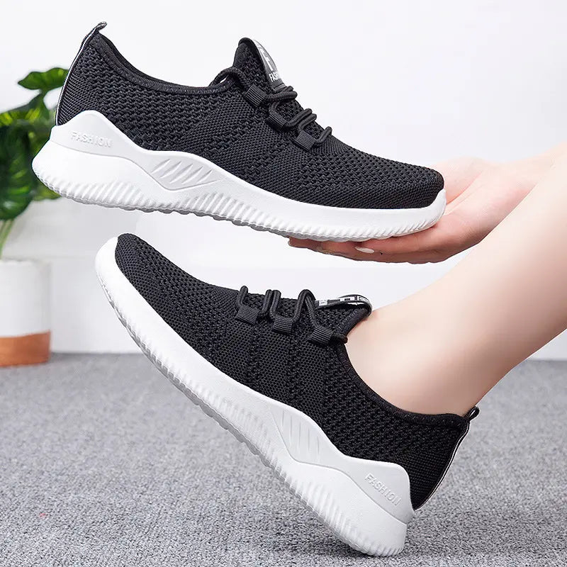 Two black Nymra women's breathable mesh trainers with white soles shown from a three-quarter angle, one held in hands and the other on a foot. The heel features subtle 'FASHION' text.