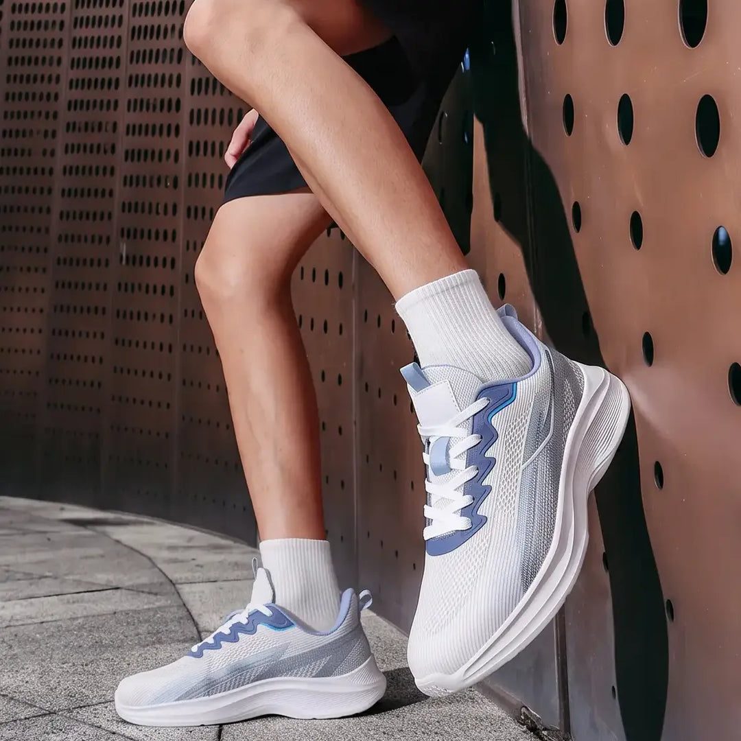 A man wearing Nyrvix lightweight breathable running shoes in steel grey with blue accents, shown in a three-quarter view leaning against an urban textured wall.
