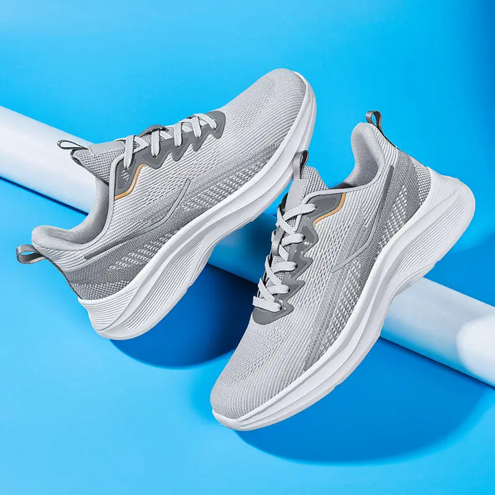 Two Nyrvix lightweight breathable running shoes in steel grey with white sculpted soles and subtle orange accents, displayed from a three-quarter angle on a vibrant blue studio background.