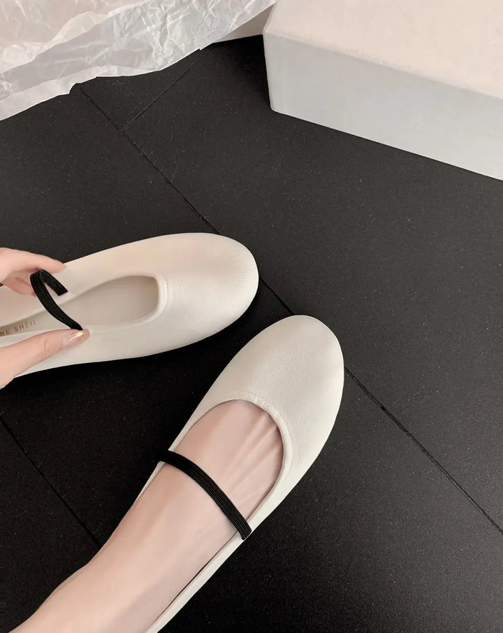 Oatmeal cream minimalist Mary Jane ballet flats with a thin black elastic strap, shown on a foot to display fit and style. The shoes have a rounded toe and a smooth matte faux leather finish.