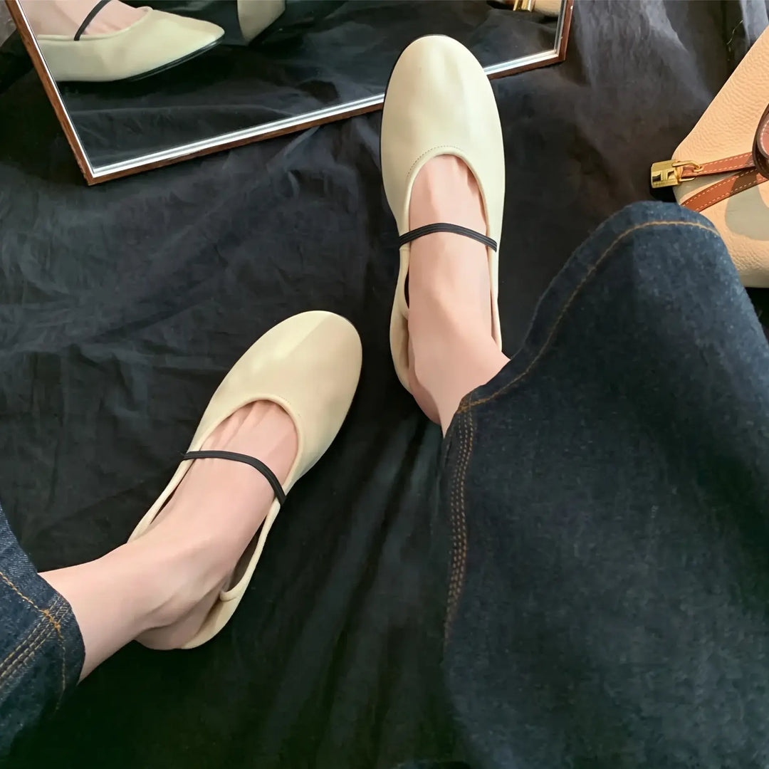 Oatmeal cream minimalist Mary Jane ballet flats with a thin black elastic strap, shown on feet with dark denim. The shoes are in matte faux leather with a rounded toe and flat sole, with a reflection in a mirror.