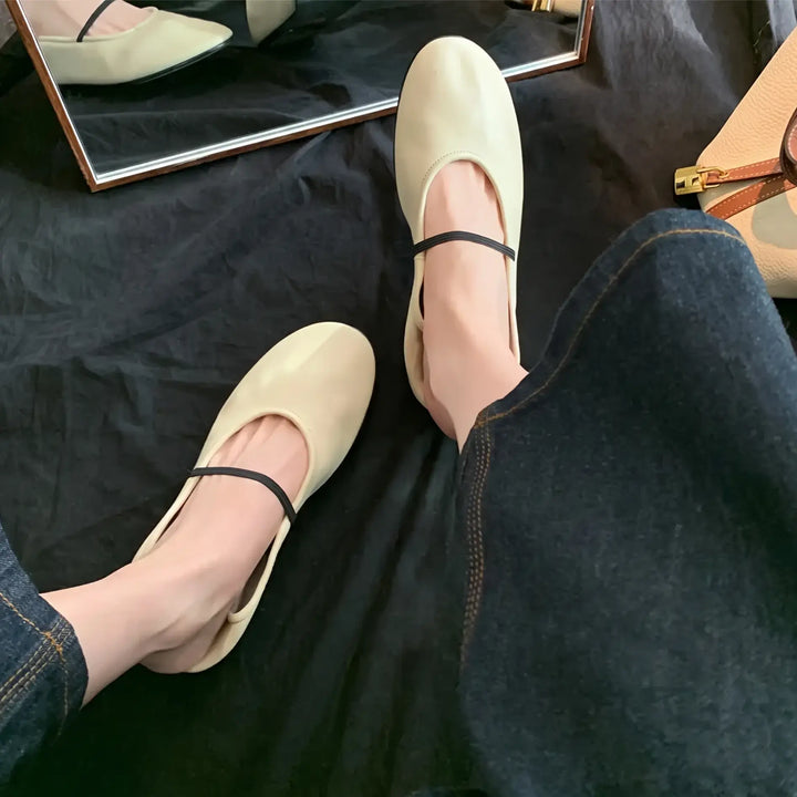 Oatmeal cream minimalist Mary Jane ballet flats with a thin black elastic strap, shown on feet with dark denim. The shoes are in matte faux leather with a rounded toe and flat sole, with a reflection in a mirror.