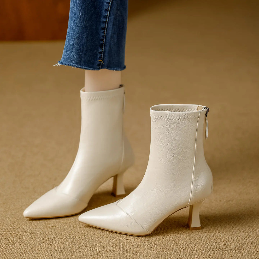 Chic off-white pointed-toe ankle boots with a back zipper and slender stiletto heel, paired with blue denim jeans in a lifestyle setting.