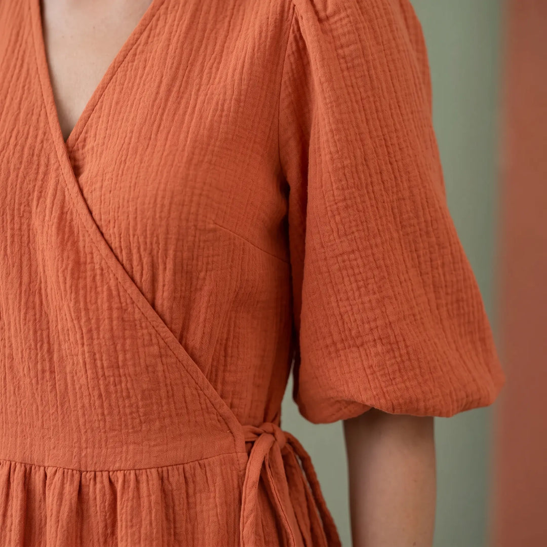 Close-up of a vibrant orange cotton gauze midi wrap dress, showcasing the crinkled fabric texture, deep V-neckline, and gathered short puff sleeve.