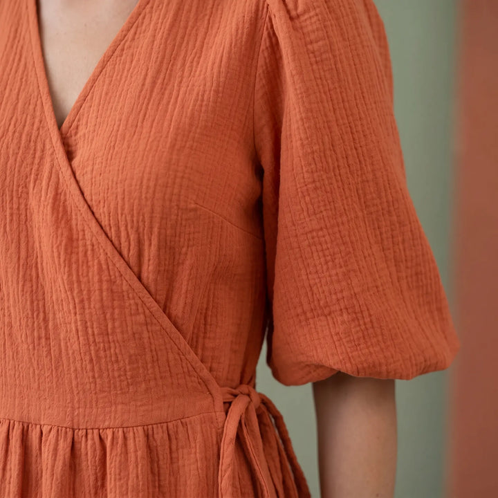 Close-up of a vibrant orange cotton gauze midi wrap dress, showcasing the crinkled fabric texture, deep V-neckline, and gathered short puff sleeve.