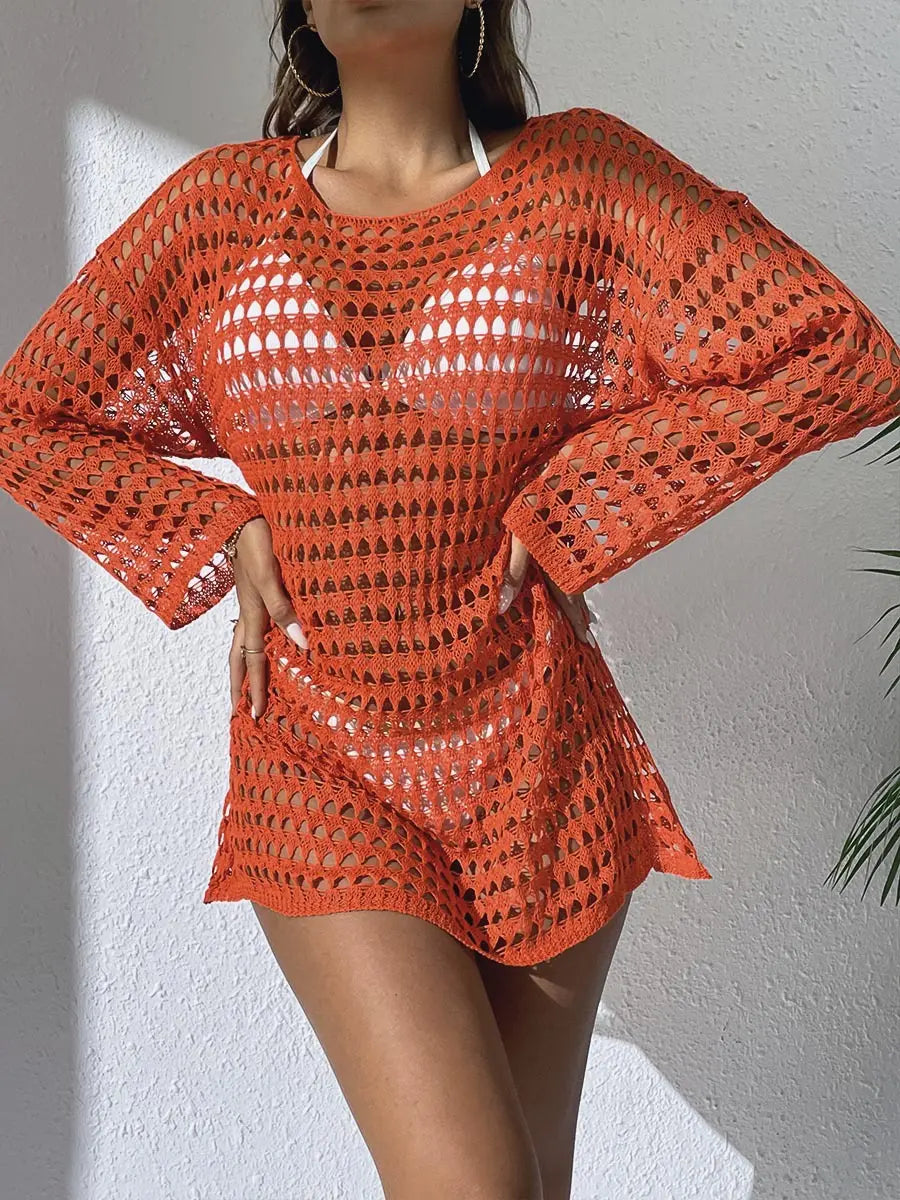 Woman wearing an orange crochet knit beach cover-up with long sleeves over white swimwear, styled in a bright resort setting. Features intricate open-knit patterns and a relaxed mini length.
