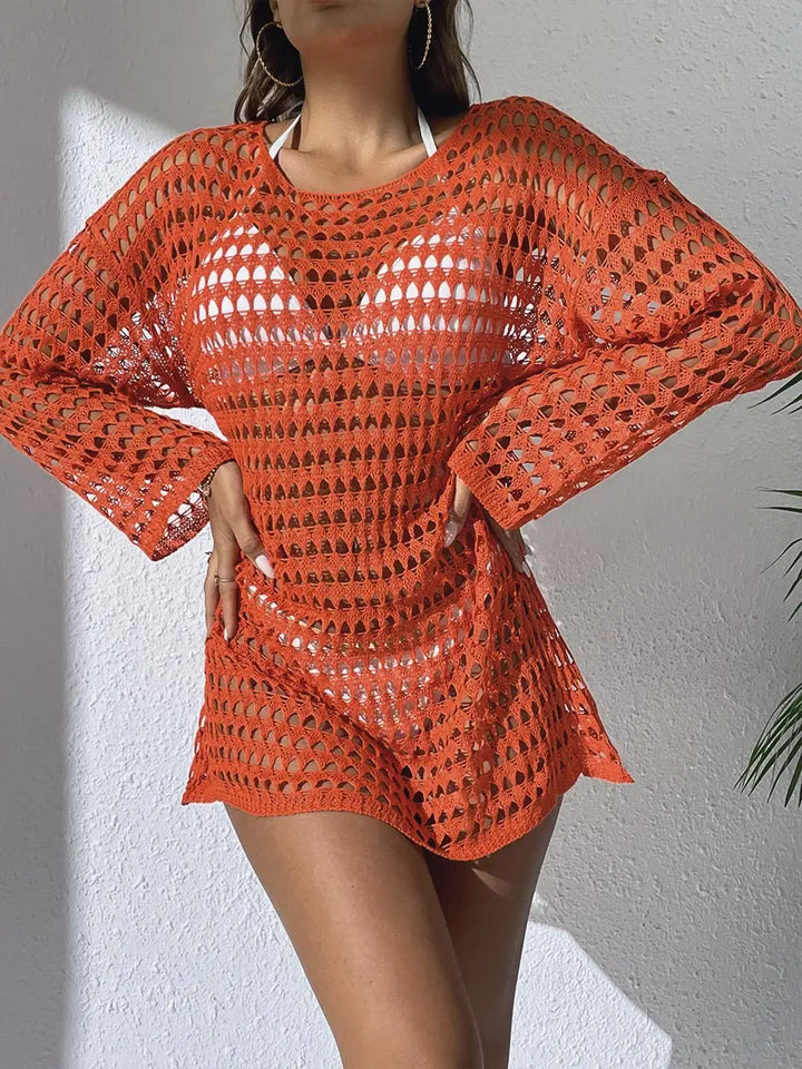 Woman wearing an orange crochet knit beach cover-up with long sleeves over white swimwear, styled in a bright resort setting. Features intricate open-knit patterns and a relaxed mini length.