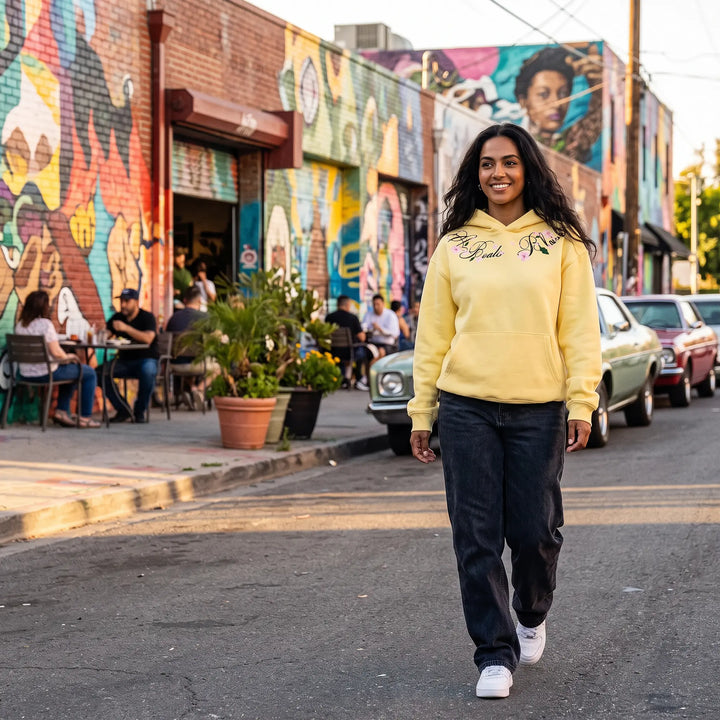 A smiling woman walks forward on a city street wearing a sunny yellow Orvela hoodie with delicate floral embroidery, dark wash jeans, and white sneakers, with vibrant murals and parked cars in the background.