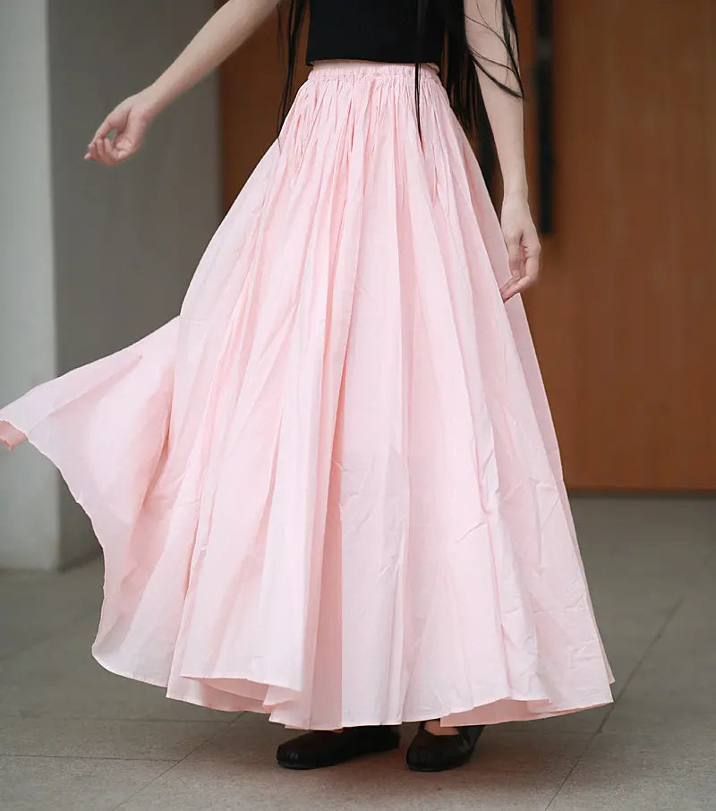 A woman models a voluminous pale pink cotton maxi skirt with a gathered elasticated waistband, showcasing its sweeping A-line silhouette and flowing fabric in an outdoor setting.