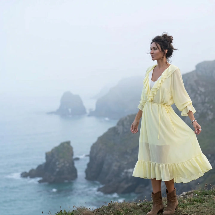 A pale yellow chiffon midi dress with cascading ruffles and a drawstring waist, shown on a model standing on a cliff overlooking the sea. The flowing, bohemian-chic dress features a V-neckline and voluminous sleeves, embodying an ethereal aesthetic.