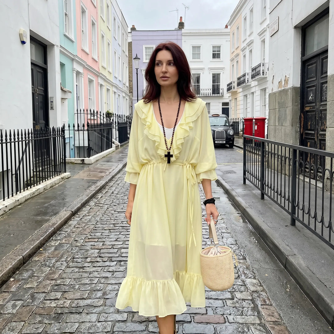 A woman walks down a charming street in a flowing pale yellow chiffon midi dress with cascading ruffles and a drawstring waist, styled for a bohemian, elegant look.