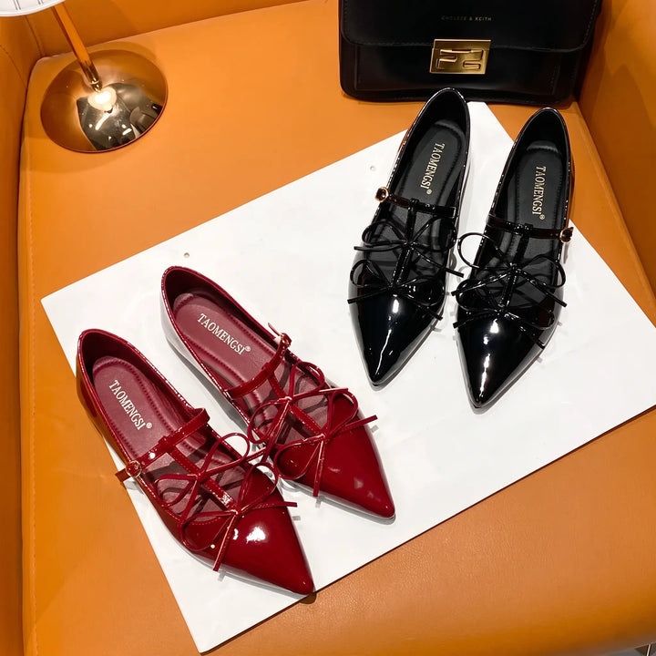 Pair of vibrant cherry red patent pointed ballet flats and classic black patent pointed ballet flats, both featuring delicate bows and adjustable ankle straps, laid out on a studio surface.