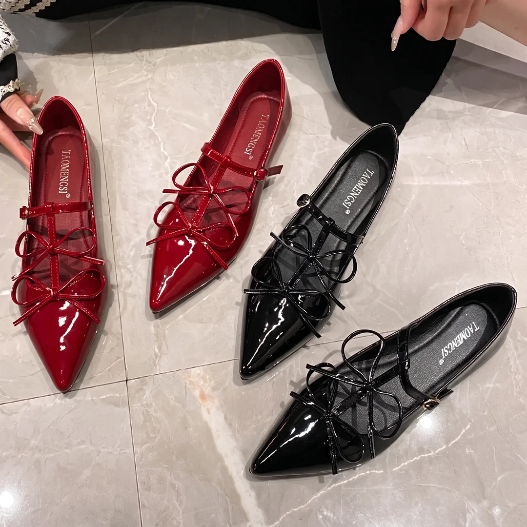 Four pairs of patent leather pointed-toe ballet flats, two in vibrant cherry red and two in classic black, featuring delicate bows and adjustable ankle straps, arranged on a marble floor.