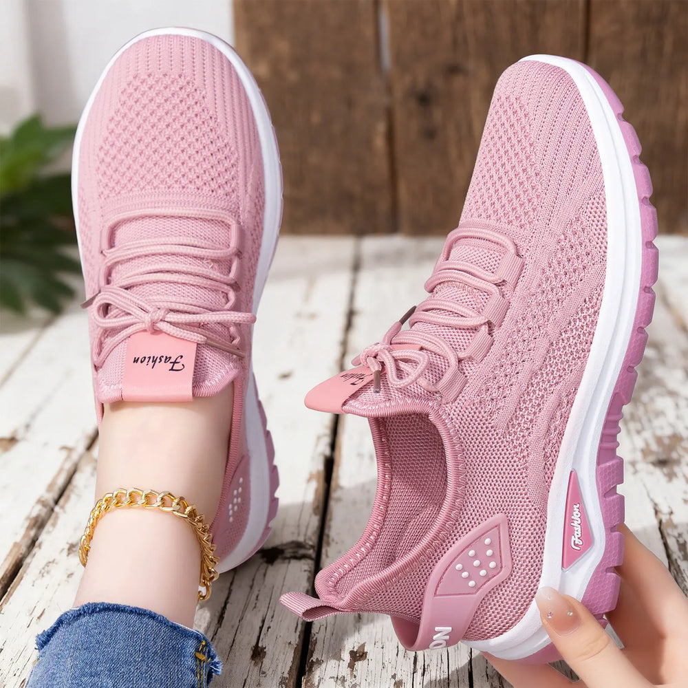 Pink breathable mesh sneakers worn on a foot with a gold anklet, alongside another shoe held by a hand against a rustic wooden background. Features white soles and 'Fashion' branding.